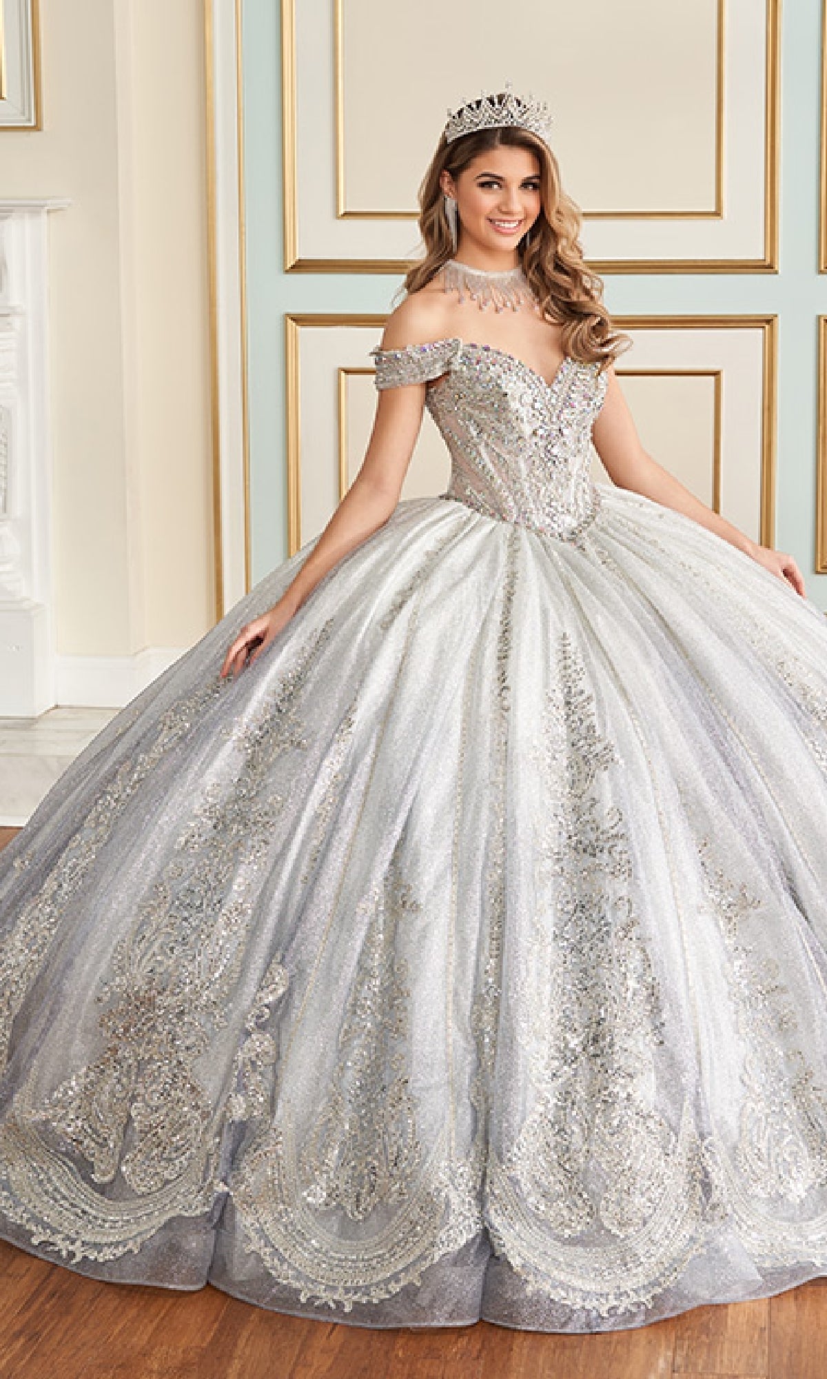 A woman in the Quinceanera Dress PR30173 By Princesa, paired with a tiara and necklace, stands indoors before a decorated wall.